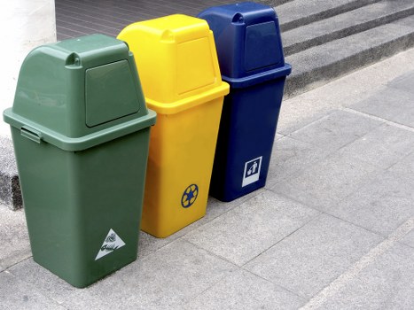 Three colour-coded outdoor rubbish bins are positioned on a paved area, likely on a driveway or public pavement near a building in Eltham. The first bin on the left is green with a smooth, matte finish and a slightly rounded, hinged lid. The middle bin is yellow, also with a matte surface and a similar rounded lid, featuring a blue recycling symbol on its front. The third bin on the right is dark blue, with a matte finish and a hinged lid, displaying a white icon of a paper or cardboard box. All three bins are upright, aligned in a neat row, and are set against a background of grey paving stones with a strip of darker textured material likely part of the pavement surface or boundary edging. The scene appears to be typical of waste management arrangements commonly seen in residential or commercial areas in Eltham, supported subtly by the service offerings of Garden Clearance Eltham, specialising in rubbish removal. The lighting is natural, indicating a clear day, and the overall setting suggests an organized and professional approach to outdoor waste storage and collection.
