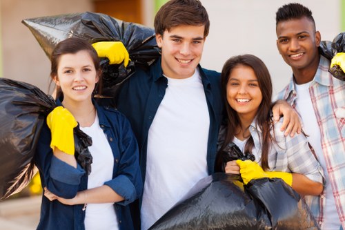 The image shows four young adults standing outdoors on a paved surface, each holding large black rubbish bags filled with waste material, which appears to be discarded household or garden waste. The group consists of two women and two men, all smiling and dressed casually; some are wearing yellow work gloves for handling rubbish. Behind them, a blurred background suggests a residential or suburban setting, potentially in Eltham, with light-colored buildings or houses. The persons are standing close together, facing the camera, with the rubbish bags resting easily on their shoulders or held in front of them. The scene conveys community involvement in rubbish collection or clearance activities, highlighting the professional waste management services provided by Garden Clearance Eltham in the local area. The overall atmosphere is positive and productive, emphasizing the importance of proper rubbish disposal and local service expertise in the Eltham vicinity.