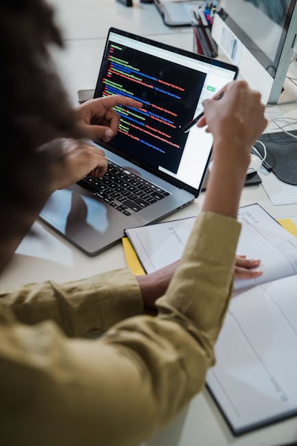 A person with curly brown hair and wearing a beige long-sleeved shirt is seated at a desk, focusing on a laptop screen displaying lines of colorful computer code on a dark background. Their left hand 