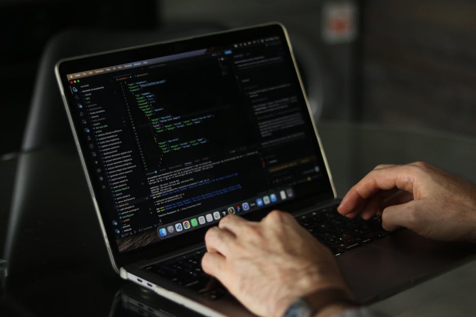 A person with curly brown hair and wearing a beige long-sleeved shirt is seated at a desk, focusing on a laptop screen displaying lines of colorful computer code on a dark background. Their left hand 