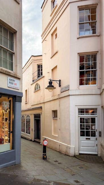 A narrow urban street scene showing the facade of a cream-colored, multi-story building with large, rectangular sash windows featuring multiple small panes of glass. The building’s exterior surface 