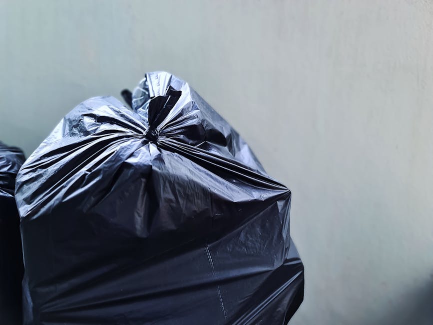 Close-up image of several black rubbish bags made of glossy plastic, tightly sealed at the top with gathered and twisted closures, positioned against a plain, light-colored wall background. The bags a
